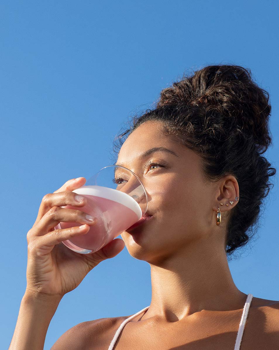 A woman standing in front of a blue sky drinking Vejo's Collagen Glow blend.