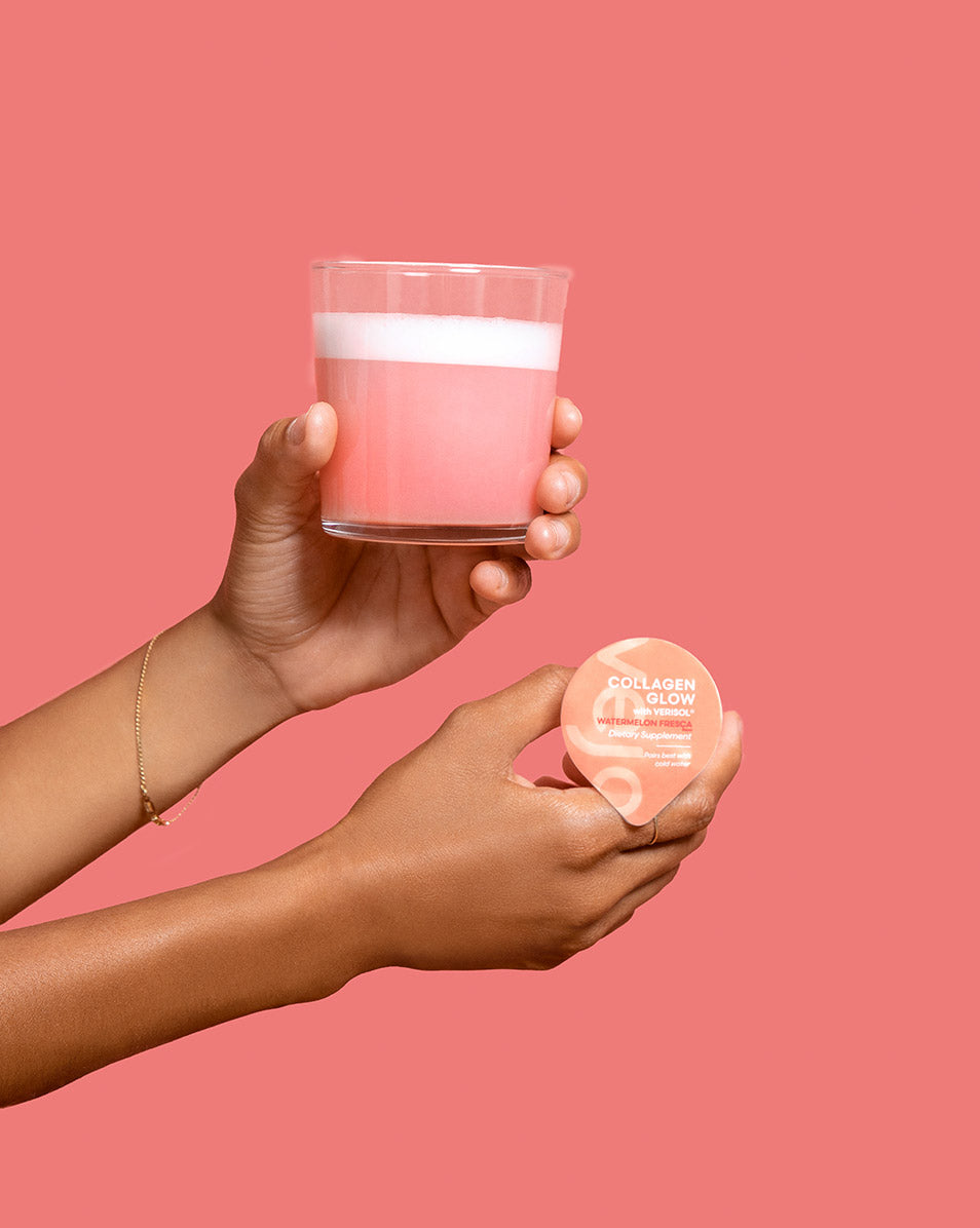 Two hands in front of a pink background, one holding Vejo's Collagen Glow blend and the other holding a Collagen Glow blending smoothie in a clear glass.