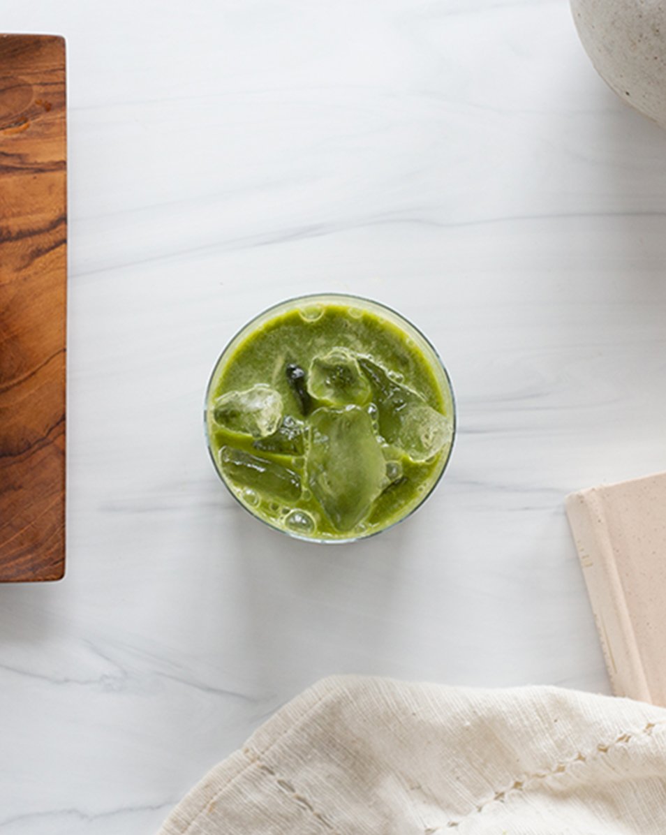 Vejo's new Matcha Latte blend photographed from overhead sitting in a glass cup with ice on a white marble countertop and kitchen towel tucked to the side of a white Vejo device.